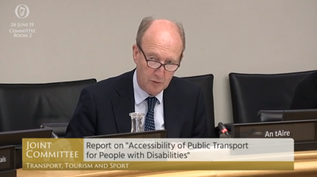Then Minister for Transport Shane Ross - wearing a dark suit jacket, light shirt and spotted tie - sitting at desk in Committee Room 2 on 29 June 2019, making the announcement of the National Transport Training Centre. His mic is lit up in red showing he is speaking. The text on the Oireachtas TV screen says - Joint Committee, Report on Accessibility of Public Transport for People with Disabilities. 