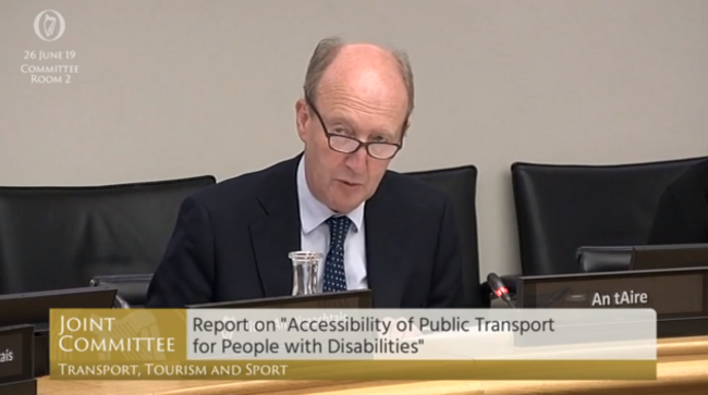 Then Minister for Transport Shane Ross - wearing a dark suit jacket, light shirt and spotted tie - sitting at desk in Committee Room 2 on 29 June 2019, making the announcement of the National Transport Training Centre. His mic is lit up in red showing he is speaking. The text on the Oireachtas TV screen says - Joint Committee, Report on Accessibility of Public Transport for People with Disabilities. 