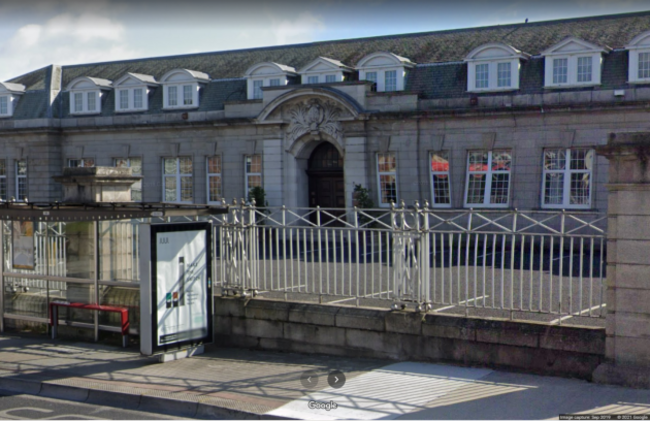 Two story building made of granite blocks with white windows and a large door surrounded by decorative stone in the middle. The windows on the second story are jutting out of the roof with a mixture of round and straight slanted roofs. There are metal railings with large piers in front of the building, with a carpark in between. A bus stop sits in front of the railings beside the road. 