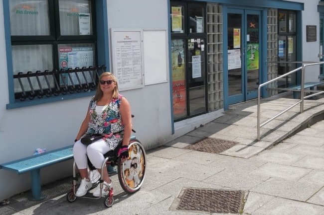 Disability activist Vicky Matthew - wheelchair user wearing sunglasses, a bright top with white trousers - outside a Bus Éireann office which has a ramp leading to its door. 