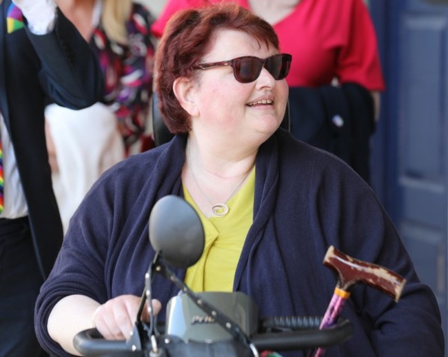 Transport activist Suzy Byrne - mobility scooter user wearing sunglasses, a yellow top and navy cardigan - with people blurred in the background. The top of Suzy’s walking stick and mobility scooter are visible. 
