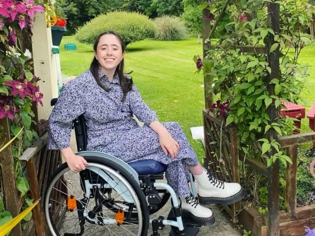 Niamh Ní Hoireahbhaird, a wheelchair user wearing a grey patterned dress and cream Dr Martens boots, in her garden beside a climbing plant with pink flowers. 