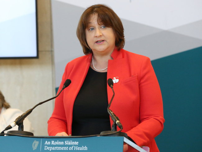 Minister Anne Rabbitte, wearing a black top and red jacket, speaking at a podium with two microphones. 