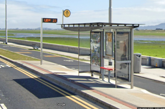 A glass bus shelter beside the sea with advertising on the back panels and a bench with a red top, with a real-time bus display on a pole nearby. The stop is located between the road and a cycle lane. There are two dished kerbs to cross the cycle lane - made of tarmac - from the bus stop to the path - both of which are made of grey concrete. There is no marked crossing on the cycle path. There is a small painted bike at the dished crossings with arrows pointed in both directions. 