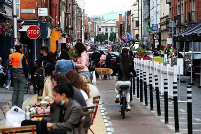 A busy street in Dublin with people sitting at tables and chairs on a path buildout that would have previously been a parking space. This has a black and white kerb at the edge, with a cycle lane running past that is protected by black and white plastic bollards. There are also people sitting at the side of a pub on chairs and standing talking near them. In the distance, there are people crossing the street, including a woman with a guide dog.