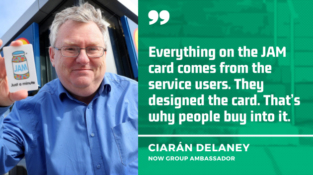 Ciarán Delaney, NOW Group Ambassador, wearing a blue shirt and holding a JAM card which has a drawing of a pot of jam and the writing ‘Just a minute’ on it, with the quote - Everything on the JAM card comes from the service users. They designed the card. That’s why people buy into it.