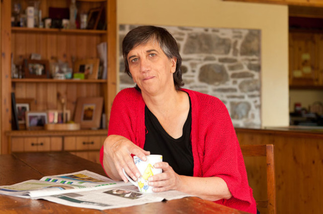 Kate Carmody wearing a black top and red cardigan sits at her kitchen table with a cup of tea in her hands.