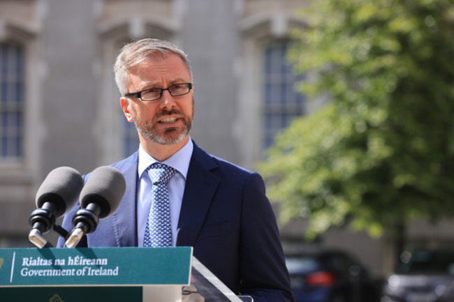 Minister Roderic O'Gorman - wearing a navy suit with light blue shirt and blue dotted tie - talking at a podium with two microphones on it. 