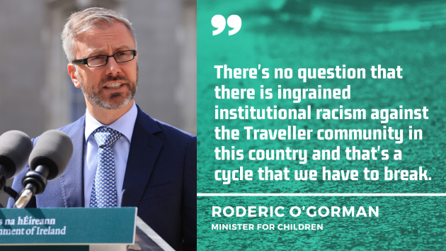 Minister Roderic O'Gorman - wearing a navy suit with light blue shirt and blue dotted tie - talking at a podium with two microphones on it. With quote:  There's no question that there is ingrained institutional racism against the Traveller community in this country and that's a cycle that we have to break.
