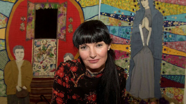 Rose Marie Maughan - wearing a red and black top - sitting in front of mural depicting a Traveller family beside a wagon