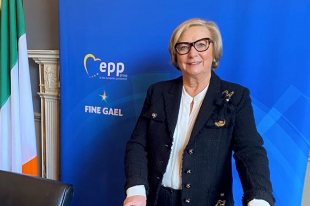 Francis Fitzgerald - wearing a white top and navy jacket with gold badges - standing at a table with a Fine Gael backdrop and an Irish flag in the corner.