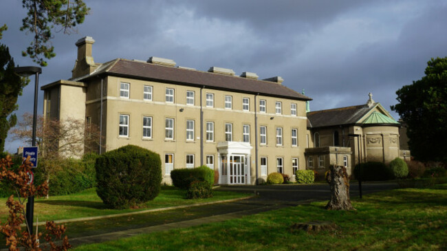 Three story grey building with many windows with white frames. The front door is surrounded by a white porch. There is a chapel adjoining the building and mature trees on the grounds.