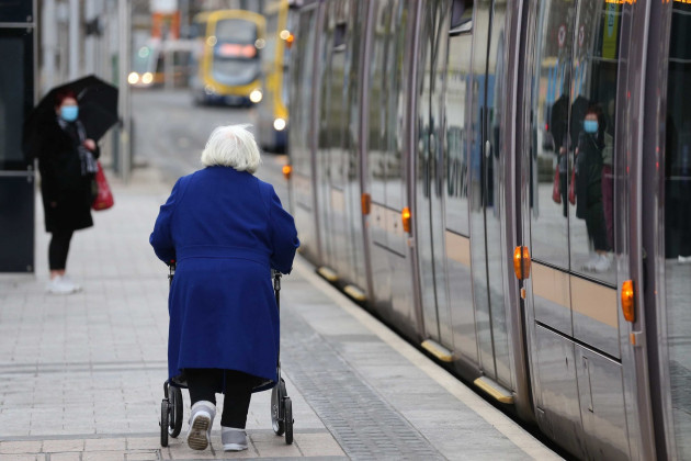 Older woman with a walker beside the Luas as it pulls into a stop in Dublin.