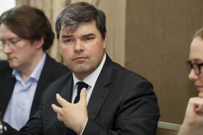 Simon McGarr wearing a white shirt with a black suit and tie, sitting in between two people at a meeting.