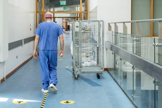Person in scrubs walking down a hospital corridor which has a mental container on wheels to its side.