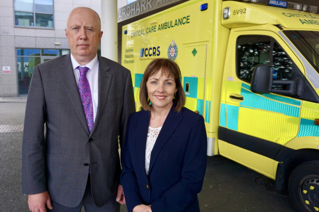 Prof Brian Lenehan - wearing a grey suit and purple tie - and Noreen Spillane - wearing a white top and navy suit jacket, standing in front of an ambulance at the emergency department.