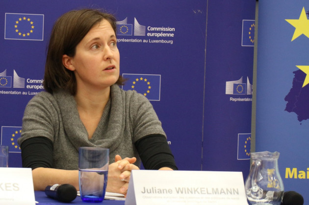 Julianne Winkelmann wearing a green top and sitting at a desk at an event with European Commission signs in the background.