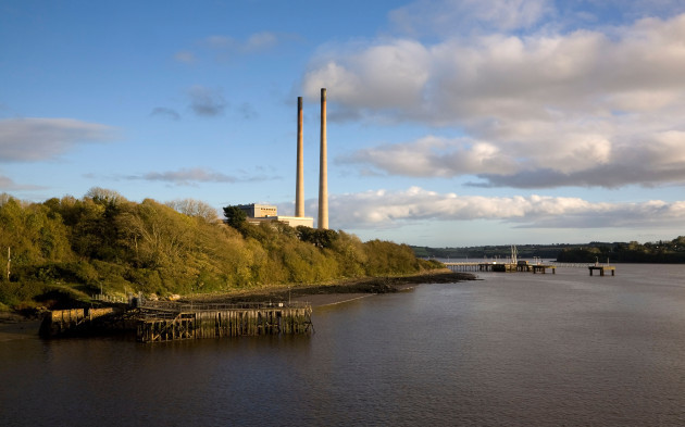 Two high chimneys stick up out of a number of trees with buildings underneath and a quay on the river beside it.