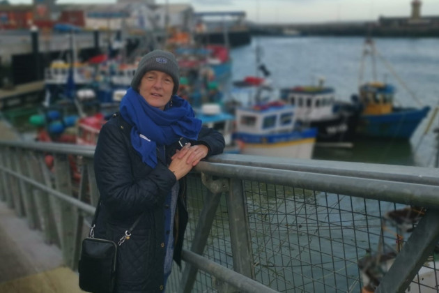 Grace O'Sullivan wearing a coat, hat and scarf standing at a pier with boats in the background.