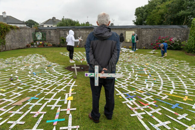 A man with grey hair and a coat stands facing away holding a white cross in his hands beside his back. Other people are in the background placing crosses. A large number of small white crosses with names form a circle on the ground. 