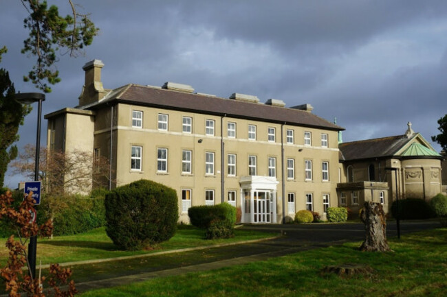 Three story grey building with many windows with white frames. The front door is surrounded by a white porch. There is a chapel adjoining the building and mature trees on the grounds.