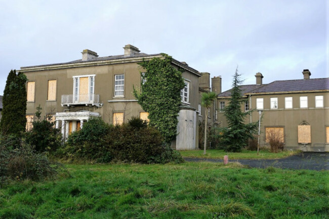 Large grey building with many windows boarded up with wooden panels. The front door is surrounded by a white porch. Other similar grey buildings are adjoined to its side with more boarded windows and doors. 