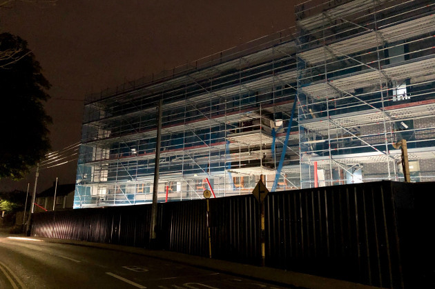A brick building with a number of floors and windows installed is visible behind scaffolding and a metal fence. It is nighttime and the construction site is lit up. 