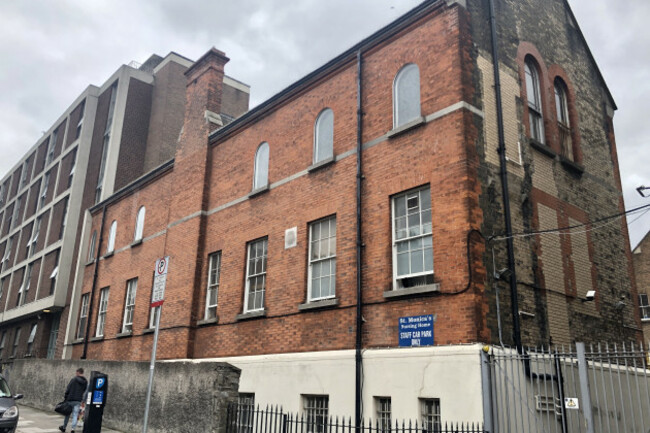 Three story building with white plastered walls on the ground floor and two red brick top floors. A sign above the ground floor says - St Monicas Nursing Home - Staff Car Park. An adjoining building which is part of the complex is a more modern building with six floors. Many of its windows are open.