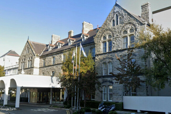 Four story grey - most likely granite - stone building with small narrow windows surrounded by carved stone. The top floor is located in the attic with small roof windows jutting out from the slate roof. A large white flat roof structure juts out from the entrance with trees and flag poles beside it.