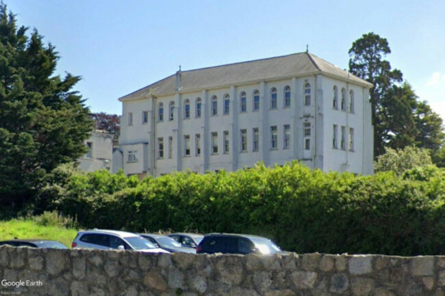 A large three story white building with many narrow tall windows and crosses on its roof is visible over a hedge with a large evergreen tree.