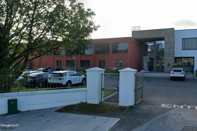 Gates leading into a carpark with the main school building behind which is a two story building with large windows and a full height glass entrance surrounded by bricks.