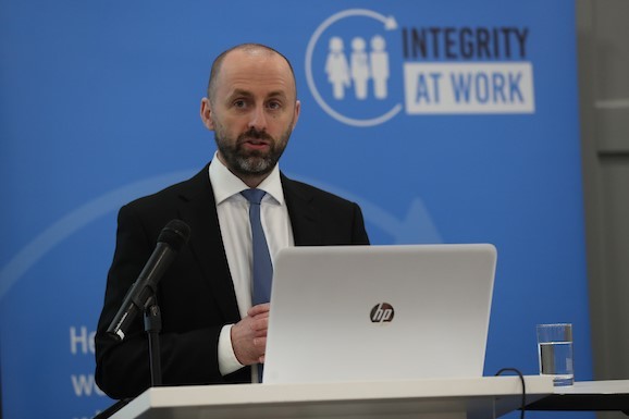 John Devitt - wearing a white shirt, suit and tie - standing at a podium speaking with the logo 'Integrity at Work' behind him.