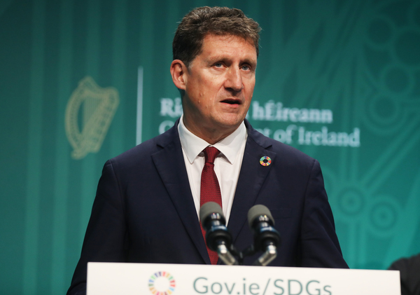Eamon Ryan speaking at a podium wearing a white shirt, red tie and dark grey jacket. The text - Rialtas nahÉireann, Government of Ireland - is blurred in the background behind him.