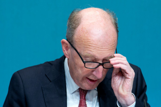 Shane Ross adjusting his glasses while looking down - wearing a white shirt, red dotted tie and dark grey suit jacket.
