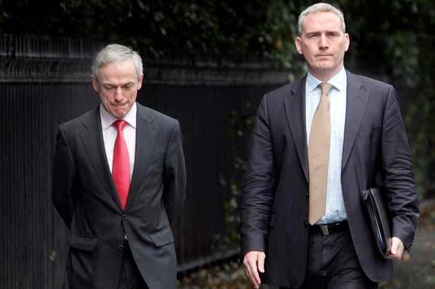 Richard Bruton - wearing a black suit with red tie - and Ciarán Conlon - wearing a dark grey suit and pink tie - are walking beside each other on a Dublin street. Conlon is holding documents in one hand.
