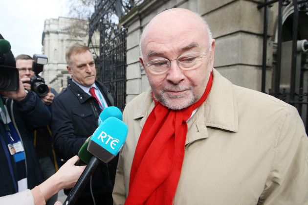 Ruairi Quinn wearing a beige coat and red scart outside the gates of Leinster House with an RTÉ and other microphones being held towards him by reporters.