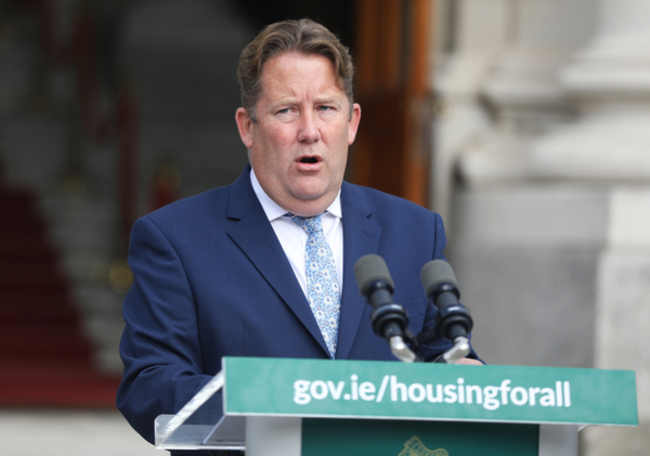 Darragh O'Brien speaking at a podium that has gov.ie/housingforall written on it. He is wearing a white shirt, light blue tie and navy suit jacket.