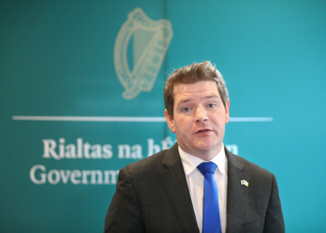 Peter Burke speaking - wearing a dark grey suit jacket and blue tie. The text - Rialtas nahÉireann, Government of Ireland - is blurred in the background behind him.