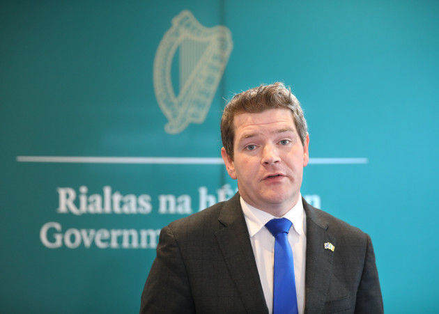 Peter Burke speaking - wearing a dark grey suit jacket and blue tie. The text - Rialtas nahÉireann, Government of Ireland - is blurred in the background behind him.