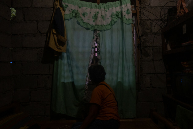 A woman sits in a room darkened to obscure her identity - in front of her window with curtain drawn.