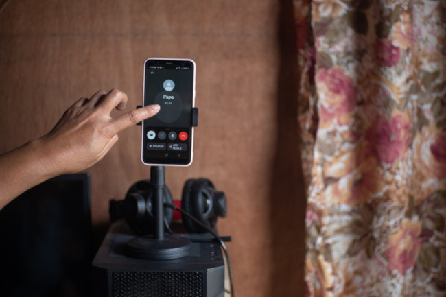 A phone is showing a call from ‘Papa’ with a person with pointed finger about to touch the point. It is located in a room with a patterned curtain.
