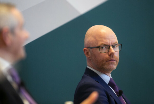 Stephen Donnelly in a suit and tie sitting at a briefing looking towards Paul Reid who is blurred in the foreground