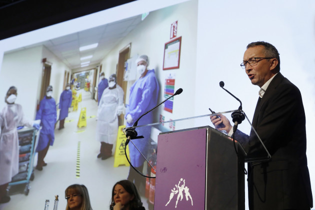 Dr Colm Henry wearing a suit and speaking a podium with an image of healthcare workers during Covid in the background