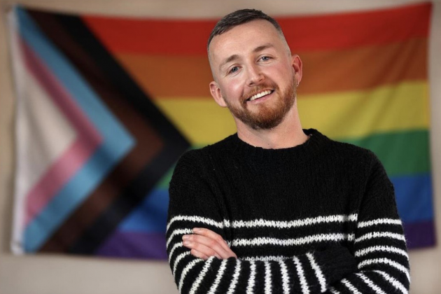 Adam Shanley wearing a black jumper with white stripes standing in front of a pride flag