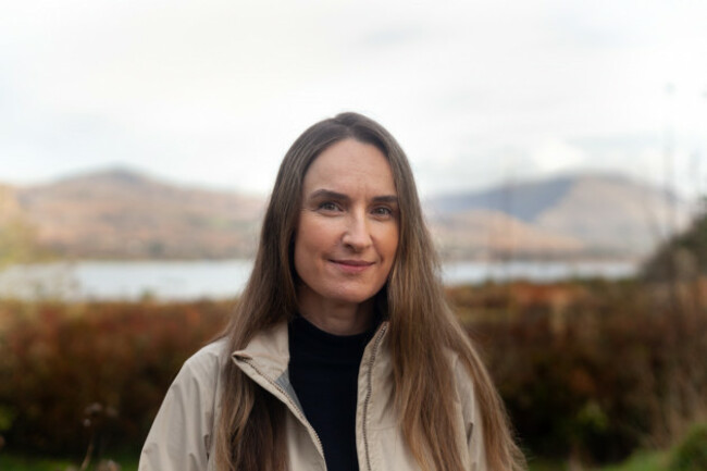 Ali Curry smiling in a grey jacket and black jumper with the sea and mountains int eh background