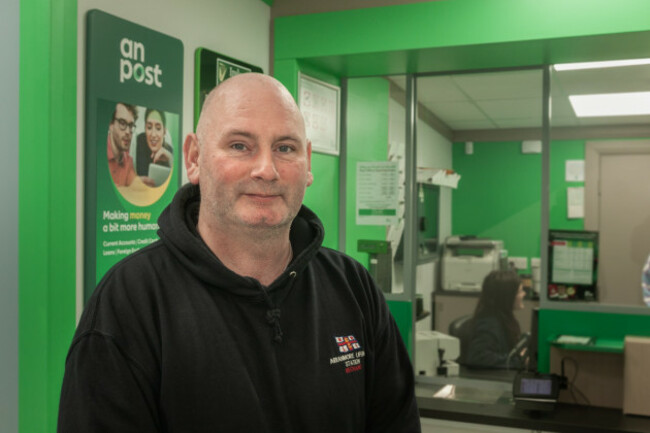 Smiling John McCafferty in a black hoodie with a post office desk in the background