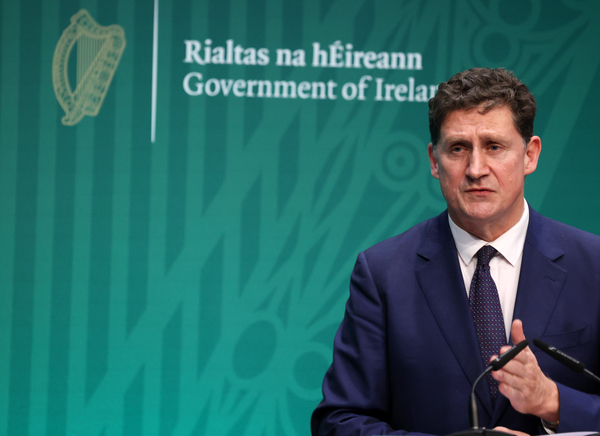 Eamon Ryan wearing a suit and tie speaking in front of a Rialtas na hÉireann Government of Ireland sign.