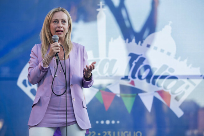 Giorgia Meloni wearing a jacket and standing in front of a sign that ends with Italia with a microphone. 