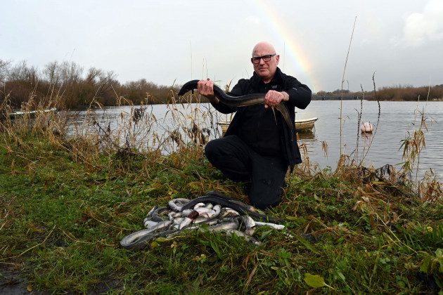 O’Connor wearing dark trousers and top hunkering on the side of the river holding a dead eel with both hands. A number of dead eels are also lying in front of him on the river bank. A small boat and rainbow can be seen in the background. 