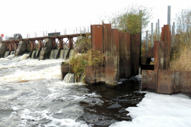 A large structure across a river made of metal that appears to be rusted as well as concrete. A gap is on the right with steps behind.